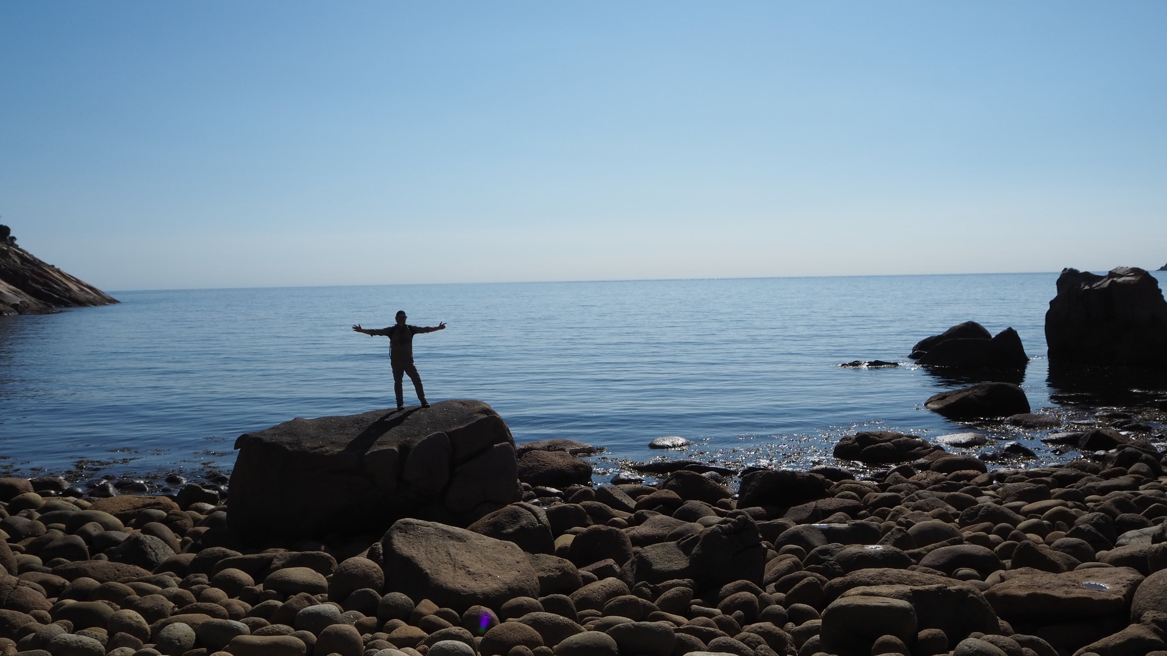 Person standing on a rock by the ocean with arms outstretched against a clear blue sky.